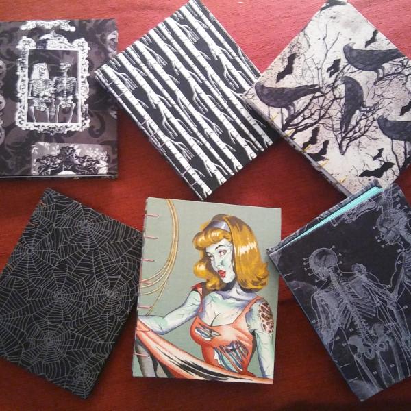 A photo of some recent Coptic bound books. From upper left: Victorian skulls, black and white trees, bats & ravens, cobwebs, zombie pinup girl, and x-ray skeleton.